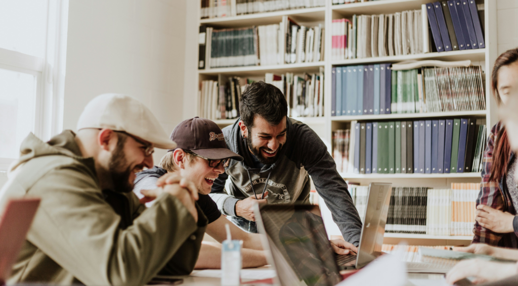 A group of young professionals gathered around a laptop, smiling and collaborating in a bright, book-filled workspace—capturing the energy of a productive, engaged team.