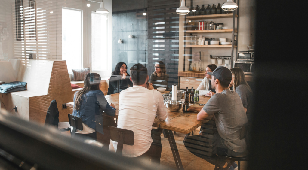 Casual team meeting in a modern office kitchen, with people gathered around a wooden table engaged in discussion and sharing drinks.