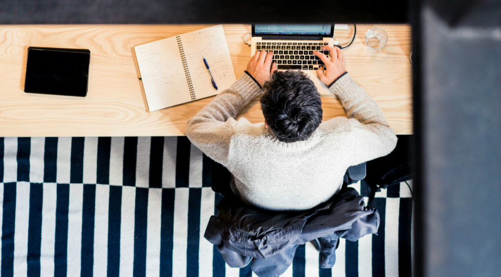 Overhead view of a person working at a wooden desk with a laptop, notebook, pen, tablet, and glass of water, seated on a striped black-and-white rug.