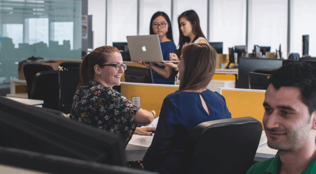 A group of coworkers collaborating in a modern open-plan office, with two women discussing at a desk and two others reviewing a laptop together.