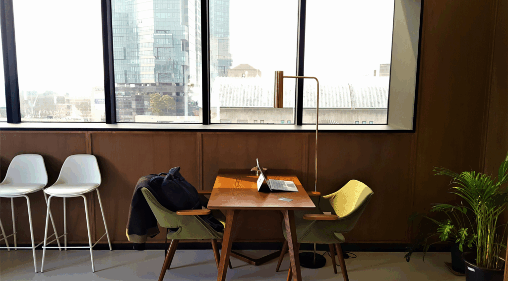 Modern coworking office space with natural light, mid-century furniture, laptop on wooden desk, and city view through large windows