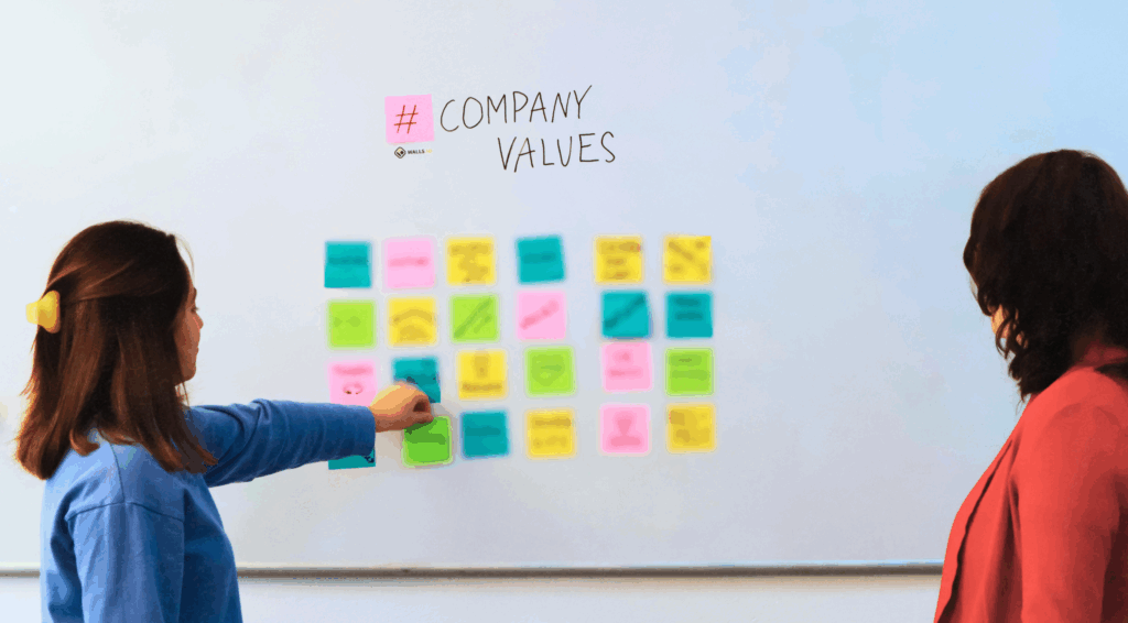 Two women stand in front of a whiteboard covered in colorful sticky notes during a company values workshop. One participant points at the notes, fostering collaboration and team alignment. The whiteboard features the words “#CompanyValues,” highlighting a culture-first approach to organizational growth.
