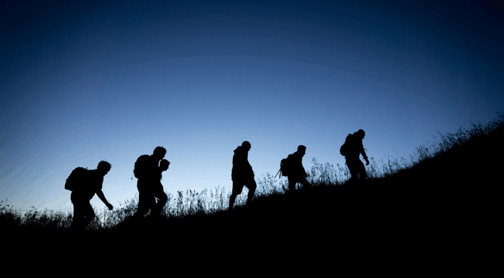Silhouette of five hikers with backpacks walking uphill at dusk against a clear blue sky.